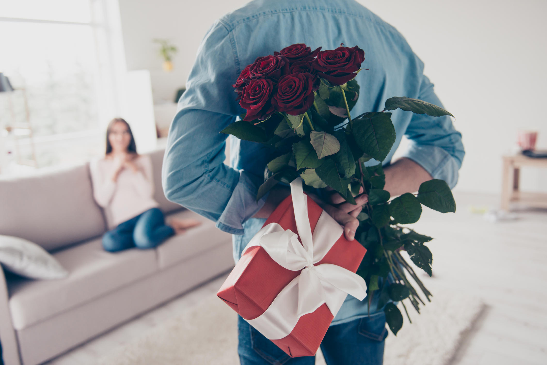 Cropped photo of man's hands hiding holding chic bouquet of red roses and gift with white ribbon behind back, happy woman is on blurred background 