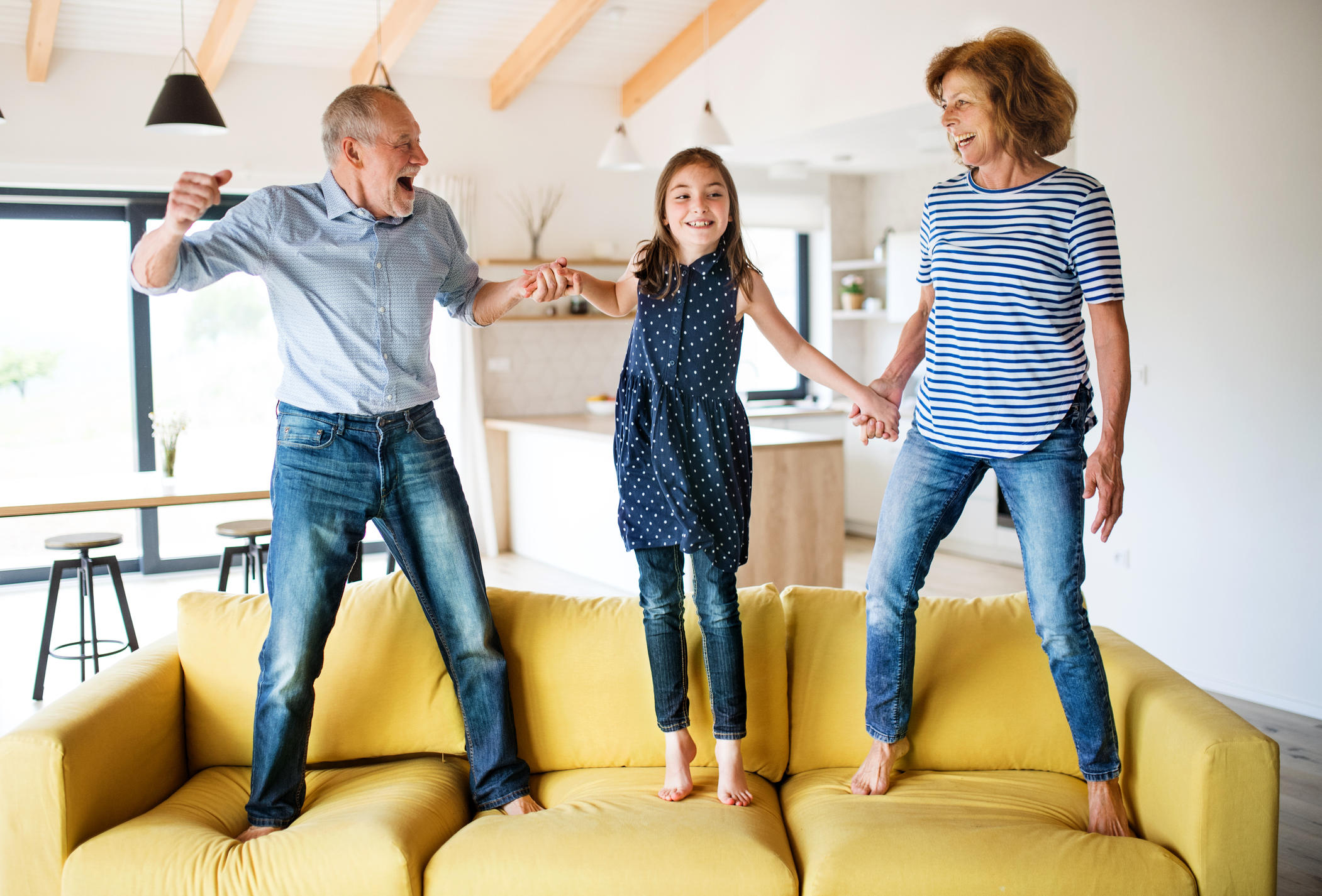 Senior grandparents and granddaughter jumping indoors on sofa, having fun. 