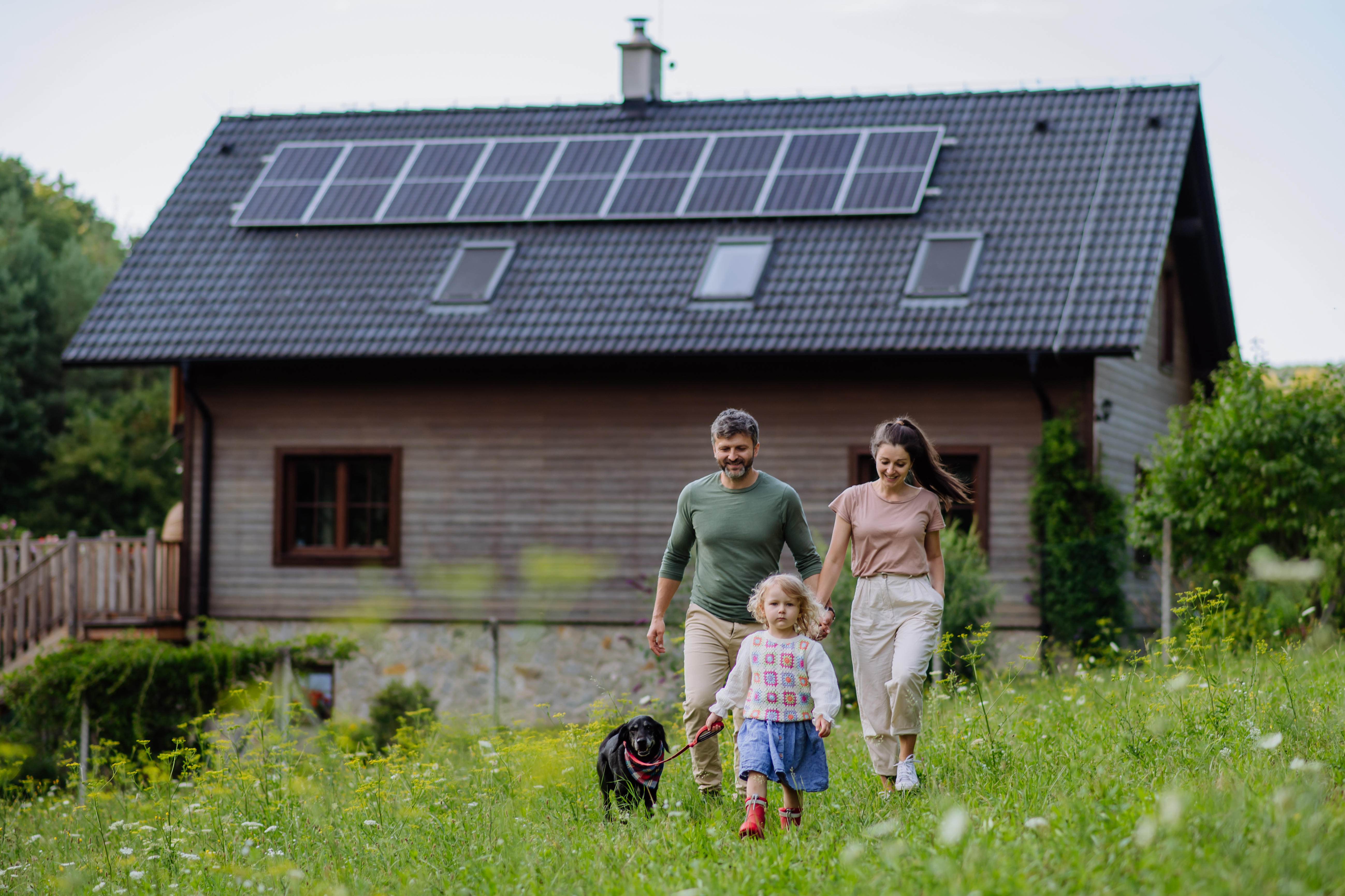 Young family with dog on a walk through the meadow near their family home with solar panels on the roof. 