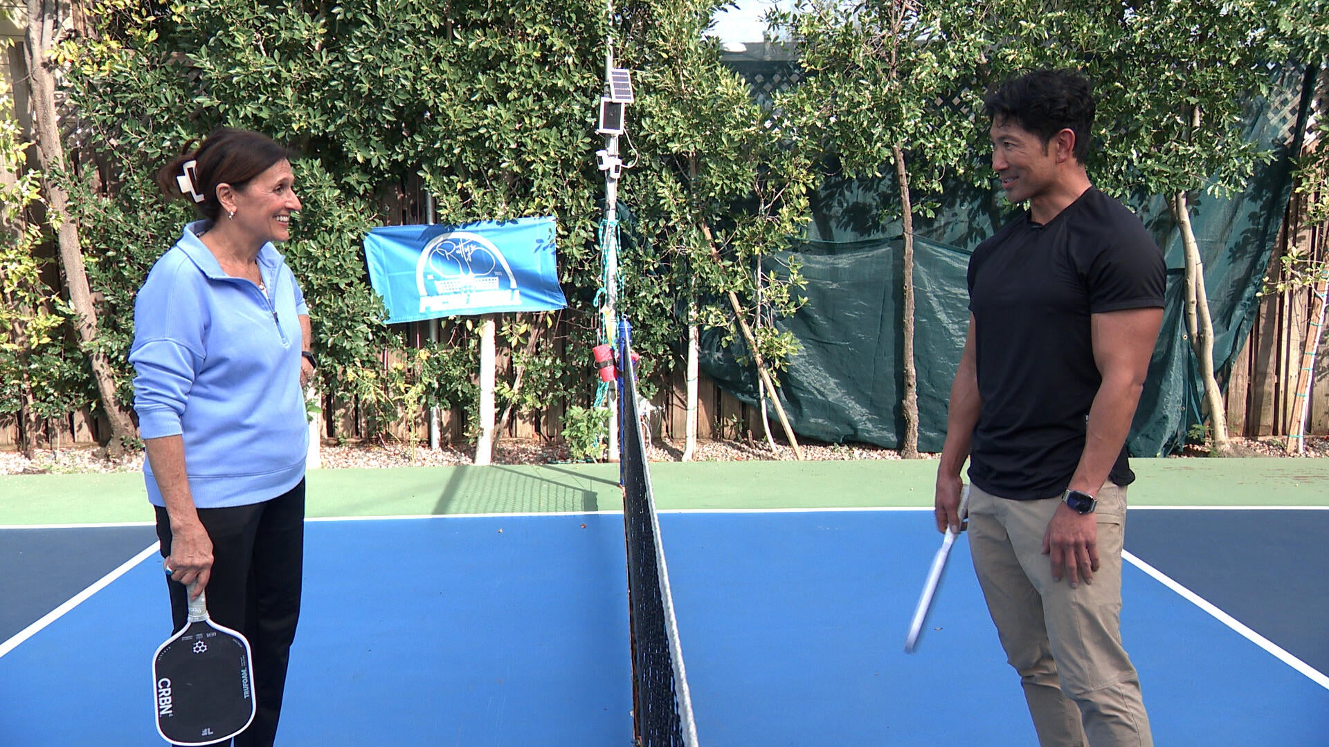 Lynn Ianni stands on the opposite side of a pickleball net from CBS News correspondent Adam Yamaguchi. qhxidiqxkiqtkinv