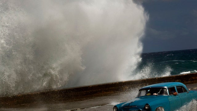 A driver maneuvers his classic American car along a wet road as a wave crashes against the Malecon in Havana, Cuba, Oct. 25, 2012.  