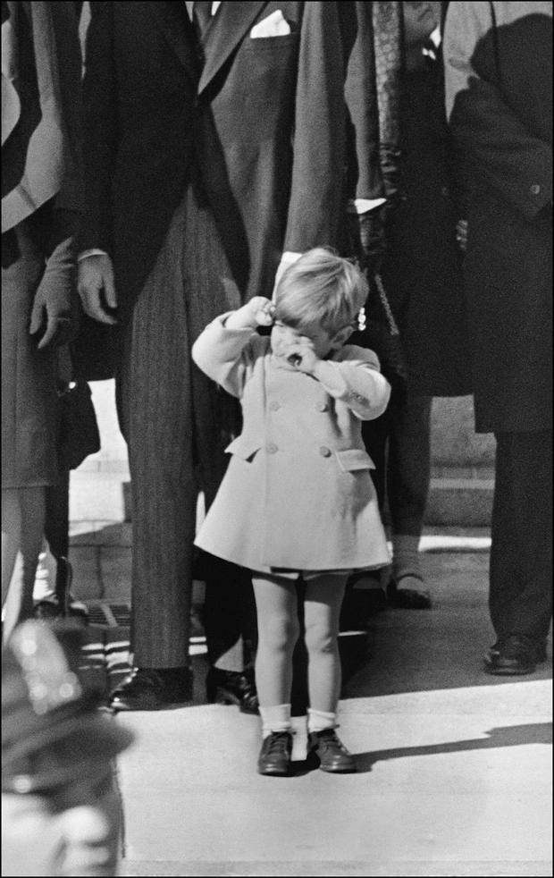 John F. Kennedy Jr wipes his eyes, near 