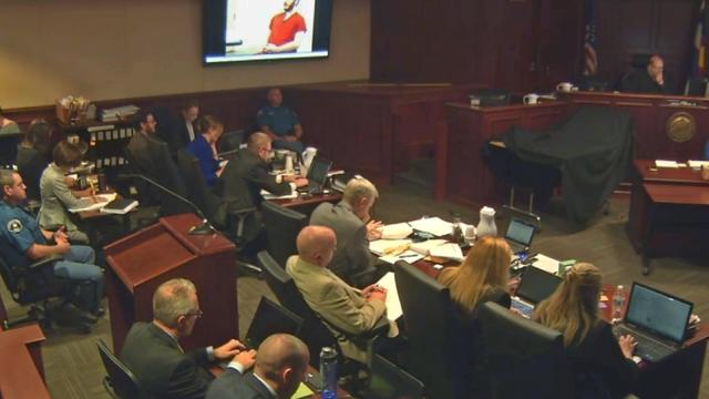 Defendant James Holmes appears in a video presented to a darkened courtroom in Centennial, Colo., June 4, 2015. Holmes, also seated at defense table below screen, spoke in the video to a psychiatrist who evaluated him for the trial on charges that he kill 