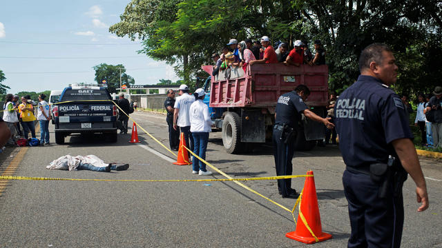 A group of migrants travelling on a lorry look at federal policemen guarding the site of an accident where a fellow immigrant died after falling off a moving vehicle during a caravan heading to the U.S., in Pijijiapan 