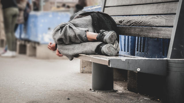 Poor homeless man or refugee sleeping on the wooden bench on the urban street in the city, social documentary concept, selective focus 