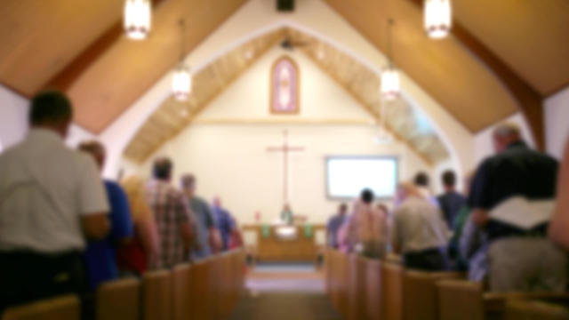 Blurred Photo of the Iterior of a Church Sanctuary with Congregation, Pastor, and a Large Cross Visible 