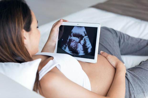 Beautiful young pregnant woman looking ultrasound of her baby on digital tablet while lying on bed. 