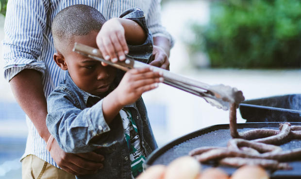 Young boy learning to use barbecue tongs with sausages 