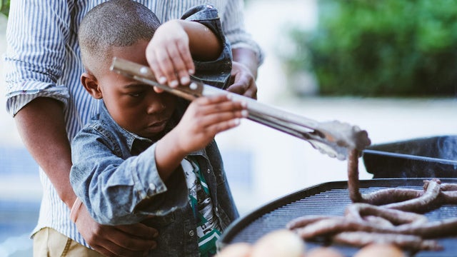 Young boy learning to use barbecue tongs with sausages 
