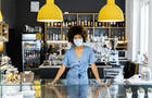 Young female owner wearing mask standing at counter in coffee shop 