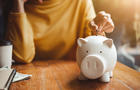 Midsection Of Woman Putting Coin In Piggy Bank On Table At Home 
