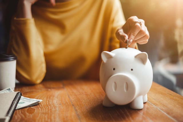 Midsection Of Woman Putting Coin In Piggy Bank On Table At Home 