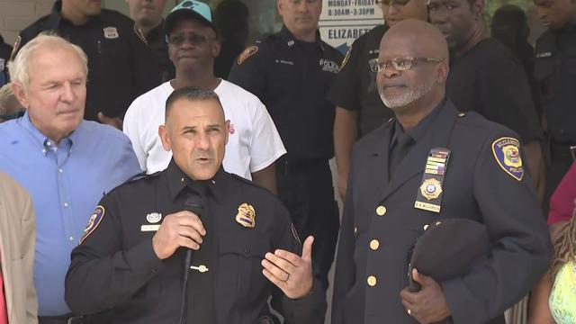 Elizabeth Police Det. Darin Williamson stands in front of a crowd of people in his police uniform, holding his hat in his arm 