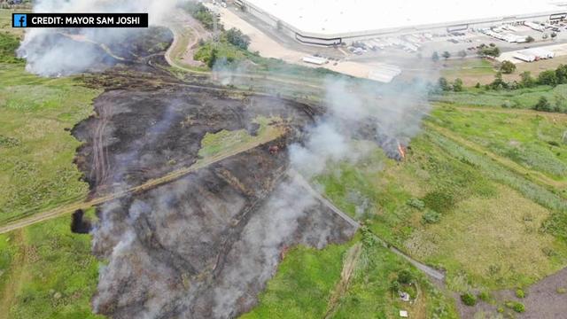 An aerial view of a green hillside with large portions of it turned gray and black from a fire and areas still smoking and burning 