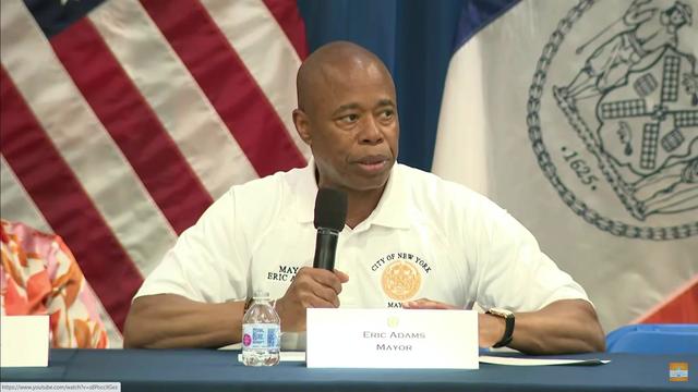 Mayor Eric Adams sits at a table in front of the American flag and the flag of New York City, holding a microphone. 