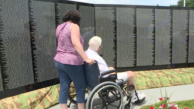A woman pushes a man in a wheelchair in front of a black wall covered with thousands of names. 