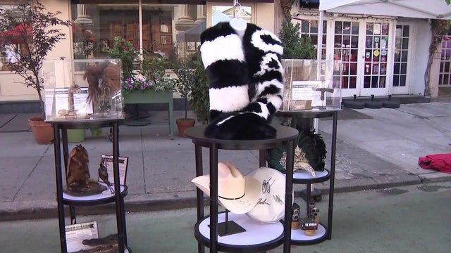 Three small tables sit in the street. On one table are a taxidermied rattlesnake, taxidermied armadillo and taxidermied crocodile head. On the middle table are a black-and-white fur, an autographed cowboy hat and what appears to be an autographed drum hea 
