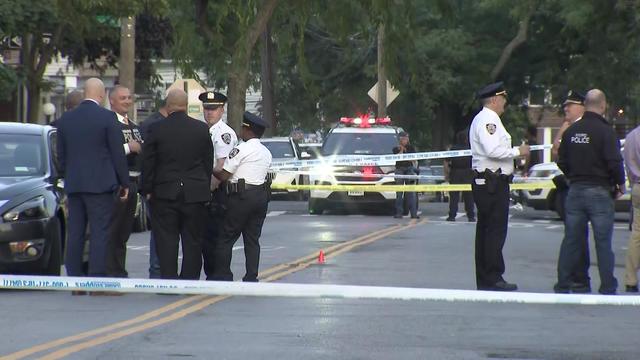 A number of police officers stand in the street between lines of police tape. An orange cone sits on the ground in the middle of the street. 