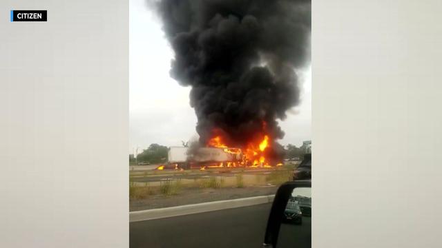 A tractor trailer on the interstate is engulfed in flames, with a large column of black smoke billowing into the sky 