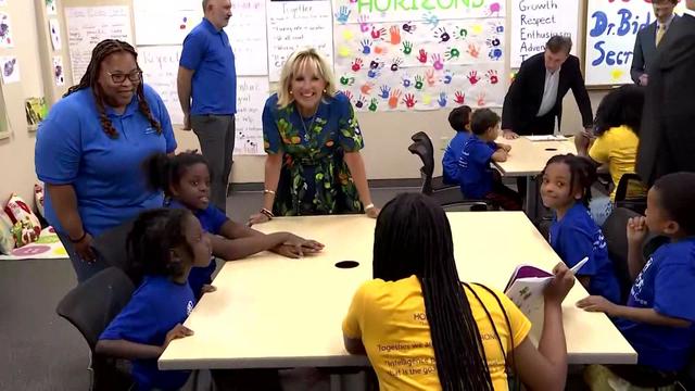 First Lady Jill Biden stands next to an instructor at a table where several students are sitting. 