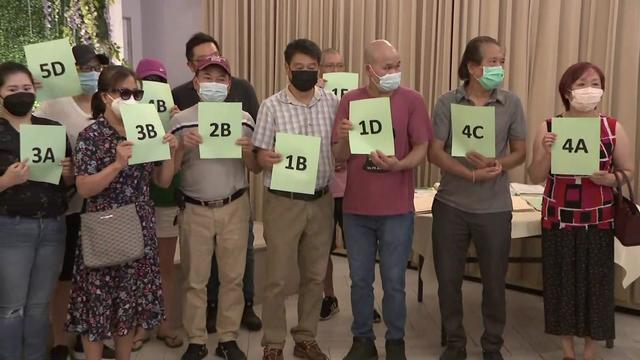 About a dozen individuals stand in a row, holding up pieces of paper showing their apartment unit number. 