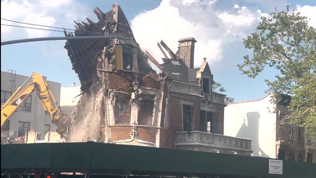 The top levels of a mansion crumble as a yellow construction crane pulls it down. 