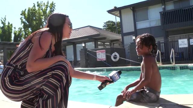 A woman with a microphone crouches at the edge of an in-ground swimming pool, speaking to a 7-year-old boy sitting with his feet in the water. 