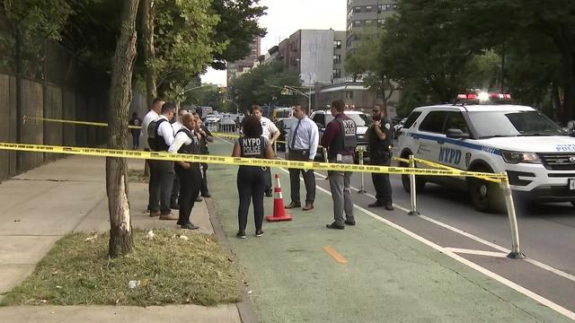 Crime scene tape blocks off a section of the sidewalk and bike lane. A group of several officers stands around an orange cone near an NYPD vehicle. 