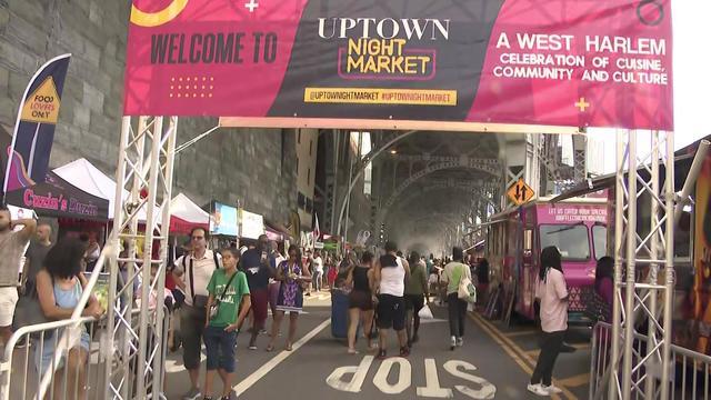 A number of booths and food trucks line the street under elevated train tracks in Harlem as crowds of people fill the street. A banner reads "Welcome to Uptown Night Market, a West Harlem celebration of cuisine, community and culture." 