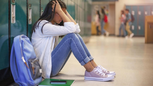 Frustrated teenage female student sitting with head in hands. Side view of high school girl in illuminated corridor. She is against metallic lockers. 