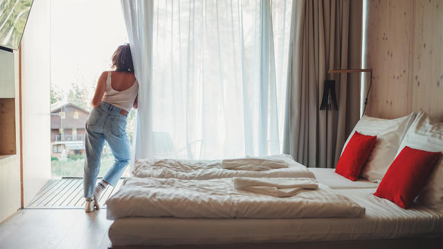 A woman dressed in blue jeans standing on forest house balcony and enjoying fresh air with nature view. Inside the Scandinavian interior design room with a king-size bed. Living in wild concept image. 