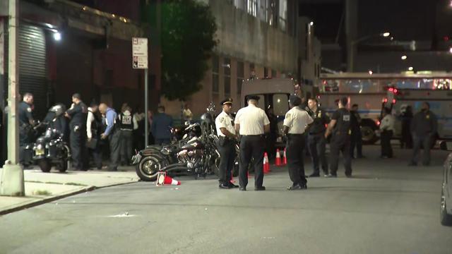 Multiple police officers stand in the street and on the sidewalk near a number of motorcycles. 