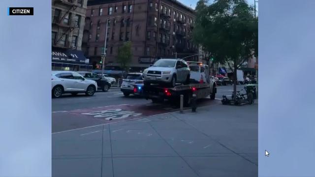 A silver Toyota sits on the back of a flatbed truck driving alongside an NYPD vehicle. 