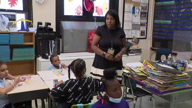 A teacher walks among desks where school children are sitting. 