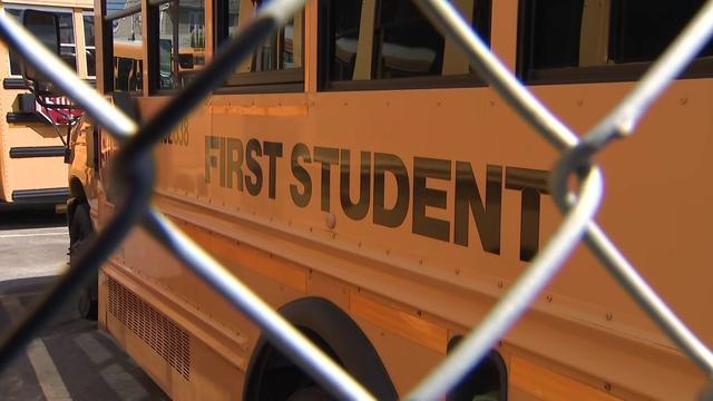 A First Student school bus sits in a parking lot behind a chain-link fence. 