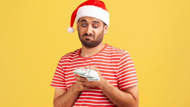 Unhappy confused bearded man in santa claus hat holding dollars in hands, greedy to buy holiday gifts 