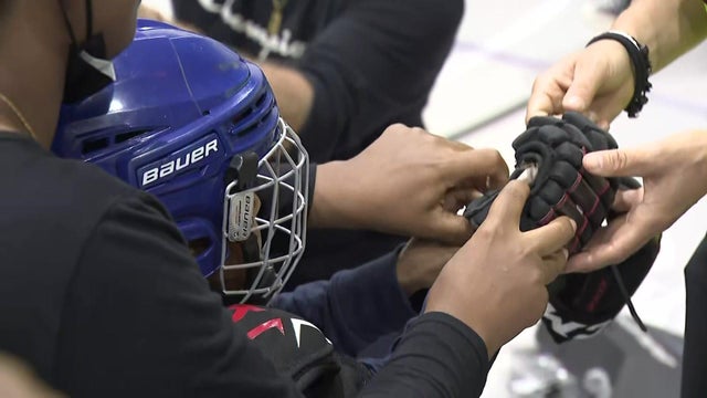 Two adults help a child in a hockey helmet put on hockey gloves. 