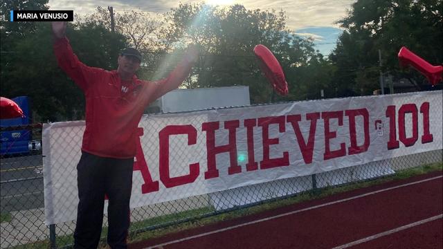 Dan O'Keefe stands on a high school track next to a banner that reads "ACHIEVED 101." 