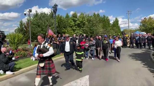 A 9-year-old boy in a firefighter's uniform marches in a small parade with other firefighters, family members and a bagpiper. 