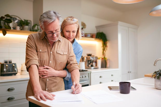 Mature Couple Reviewing And Signing Domestic Finances And Investment Paperwork In Kitchen At Home 