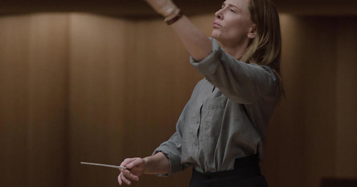 Cate Blanchett leads an orchestra in Tár with her lips pursed and her hand holding a conductor's wand. 