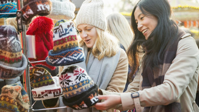 Female friends are looking at woolen hats at outdoor Christmas market stall. 