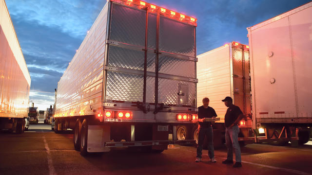 Two truck drives checking dispatch papers while standing next to truck trailers in a large parking lot at night. 