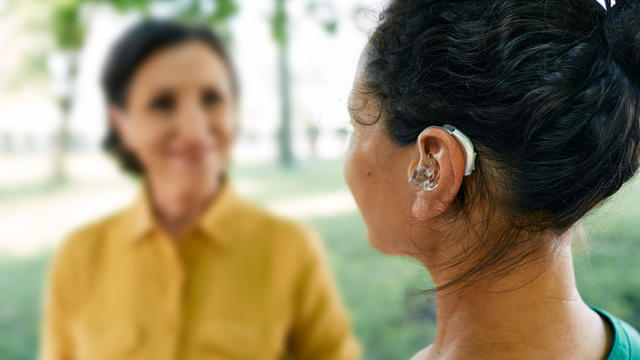 Adult woman with a hearing impairment uses a hearing aid to communicate with her female friend at city park. Hearing solutions 