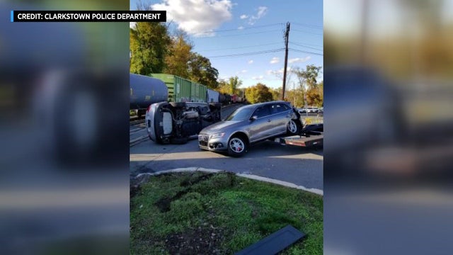 One vehicle lays on its side and another is partially off a trailer next to a freight train on train tracks. 