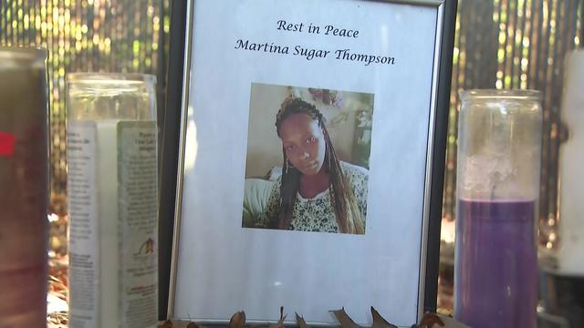 A photo of a woman with the words "Rest in Peace Martina Sugar Thompson" sits on the ground next to prayer candles. 