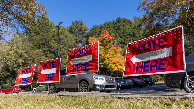 Four red "vote here" signs in front of parked cars 