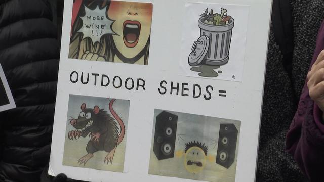 A sign at a rally that says "Outdoor sheds =" along with pictures of a rat, a full trash can, music speakers and a person yelling "more wine!!!" 