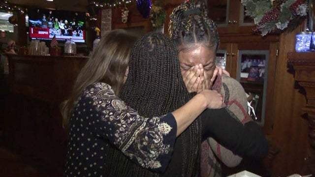 Two women hug a third woman, who is crying, after being surprised with cars and other gifts. 