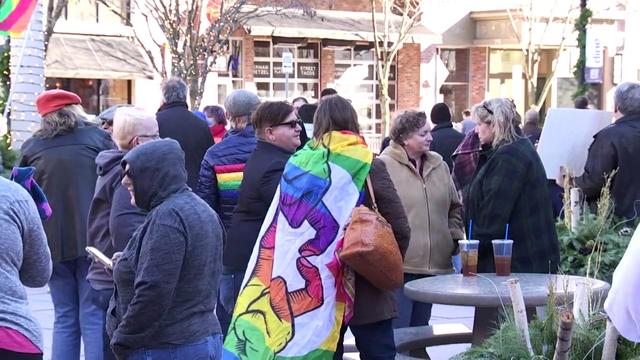 Dozens of people, some wearing or carrying rainbow items or clothing, stand in an outdoor plaza. 